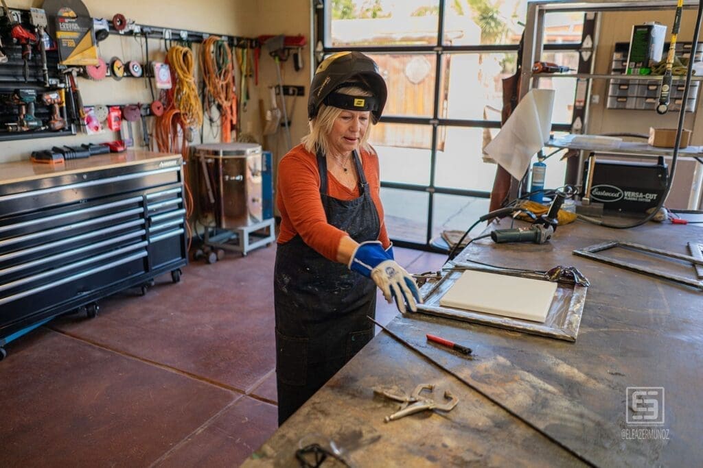 The artist looks up from a metal frame while standing at a workbench filled with tools and equipment.