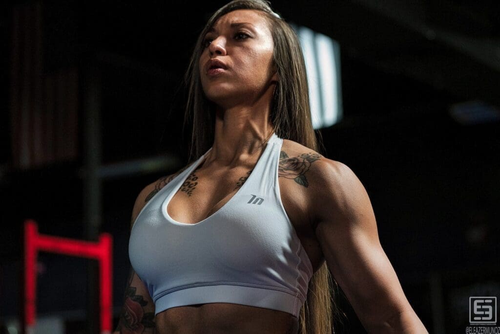 Danni looks ahead under dramatic gym lighting in a low-angle portrait that emphasizes her shoulders and upper body.
