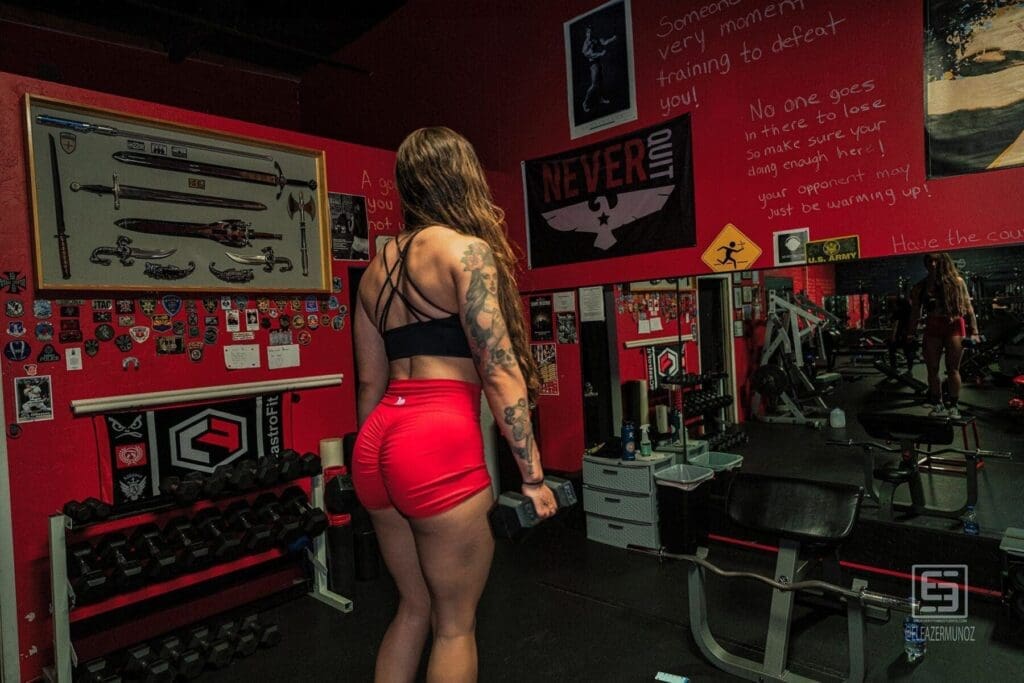 Danni stands with dumbbells beneath a wall of training notes, showing the crossed straps of her top and arm tattoo.