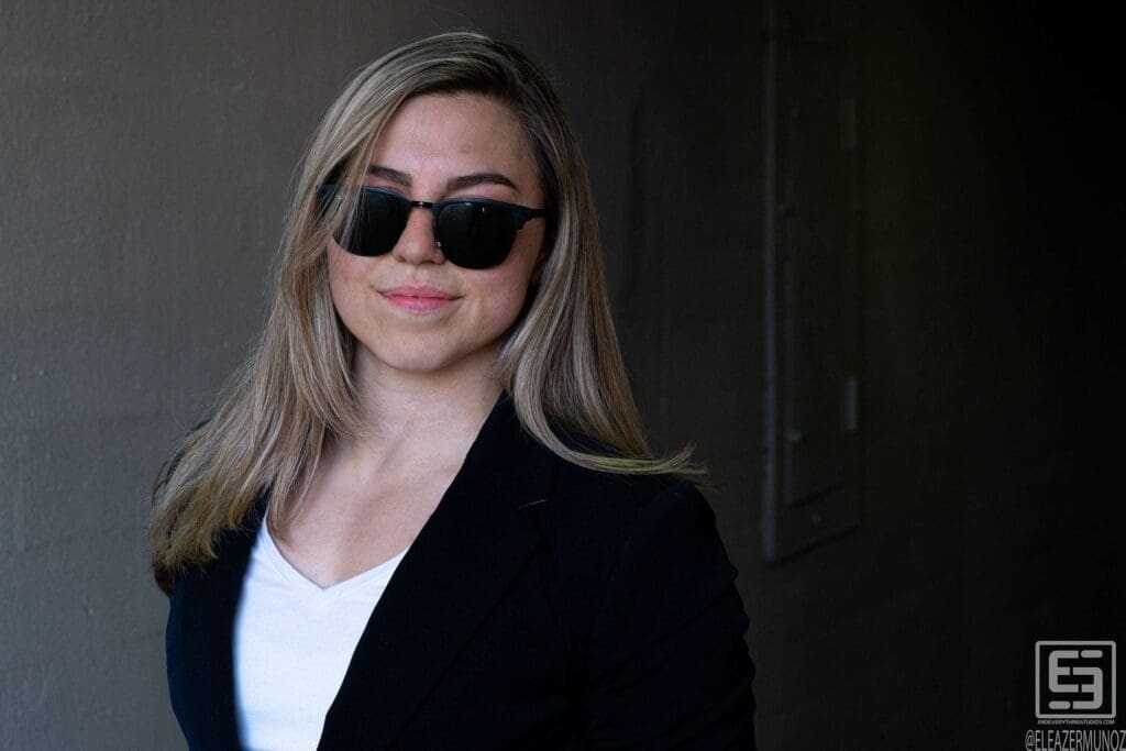Day Muñoz smiles in sunglasses and a navy blazer against a dark shaded wall.