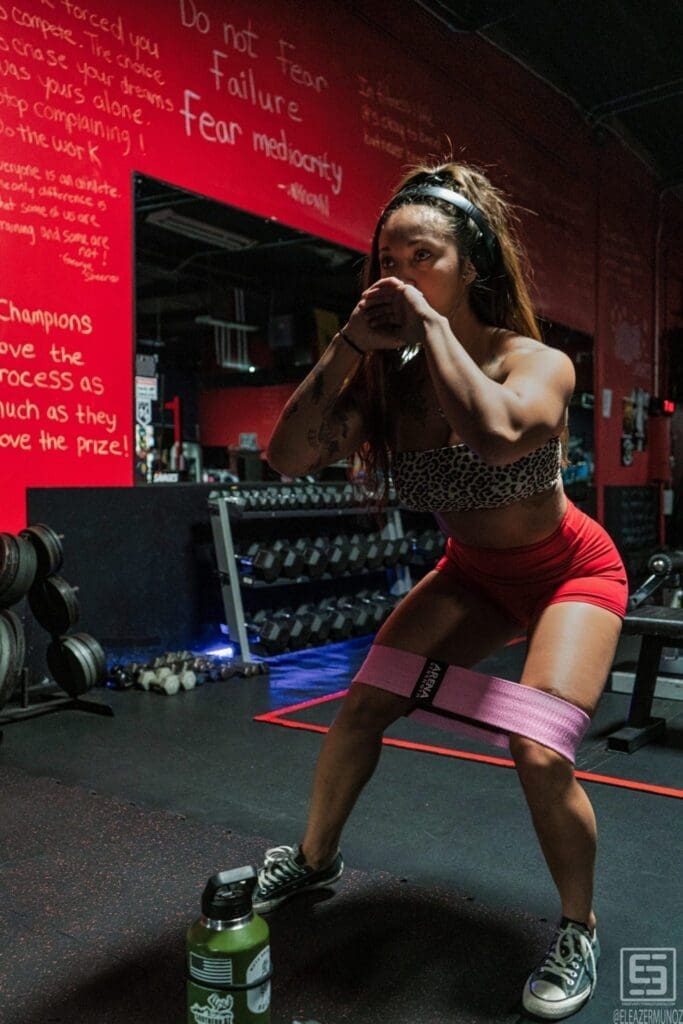 Danni holds a wide squat with a pink resistance band while looking to the side.