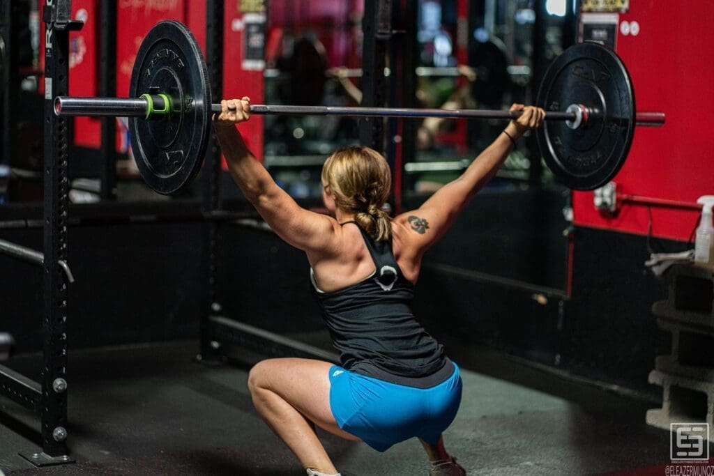 Day holds a barbell overhead in a deep squat, photographed from behind inside the rack.