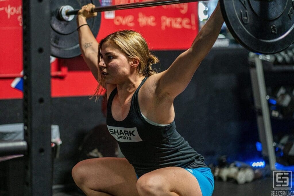 Day holds a barbell overhead while rising from a squat in a strong front-facing composition.