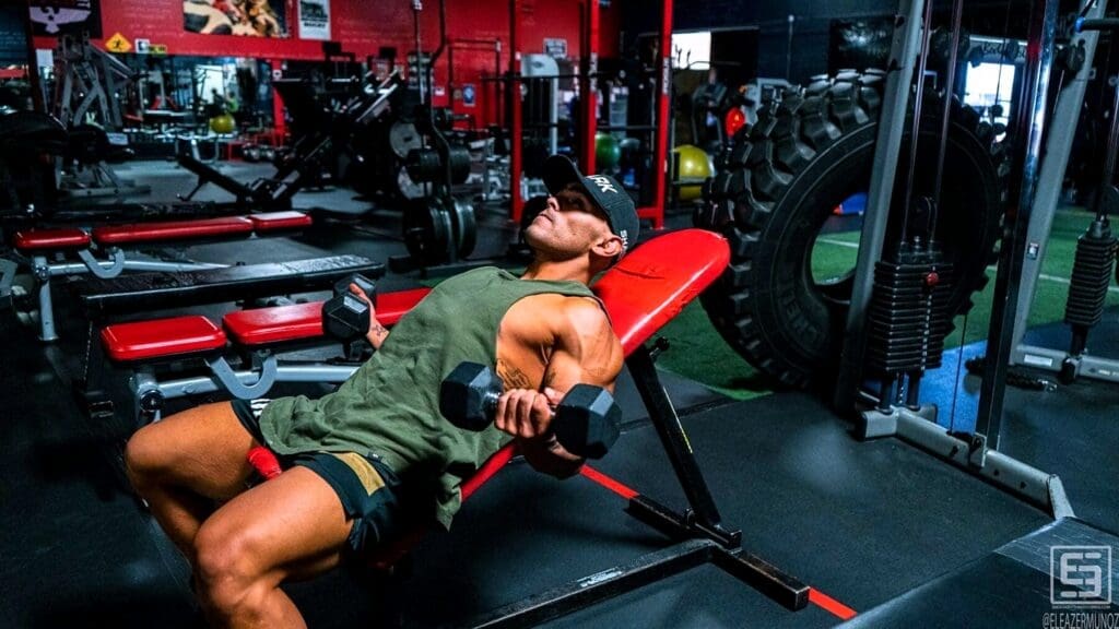 Christopher reclines on a red incline bench and curls two dumbbells in a wide gym portrait.