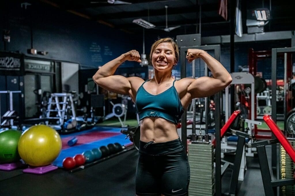 Day smiles while holding a front double-biceps pose in the gym.