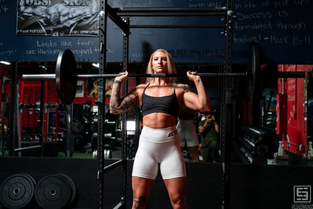 Cami Bee faces the camera with a barbell held at shoulder height inside a gym rack.