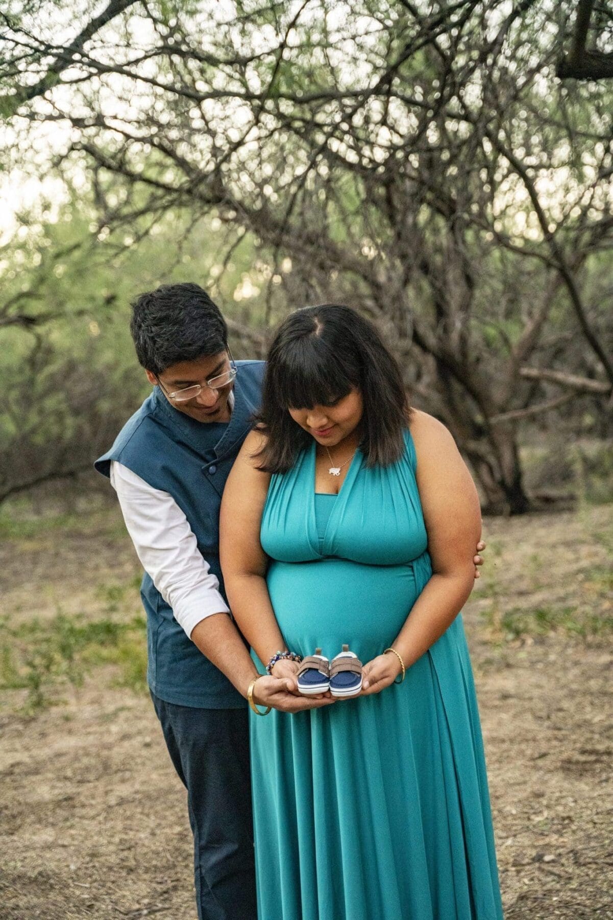 Aditi holds a pair of baby shoes over her teal maternity dress while her partner stands close behind her beneath desert trees.