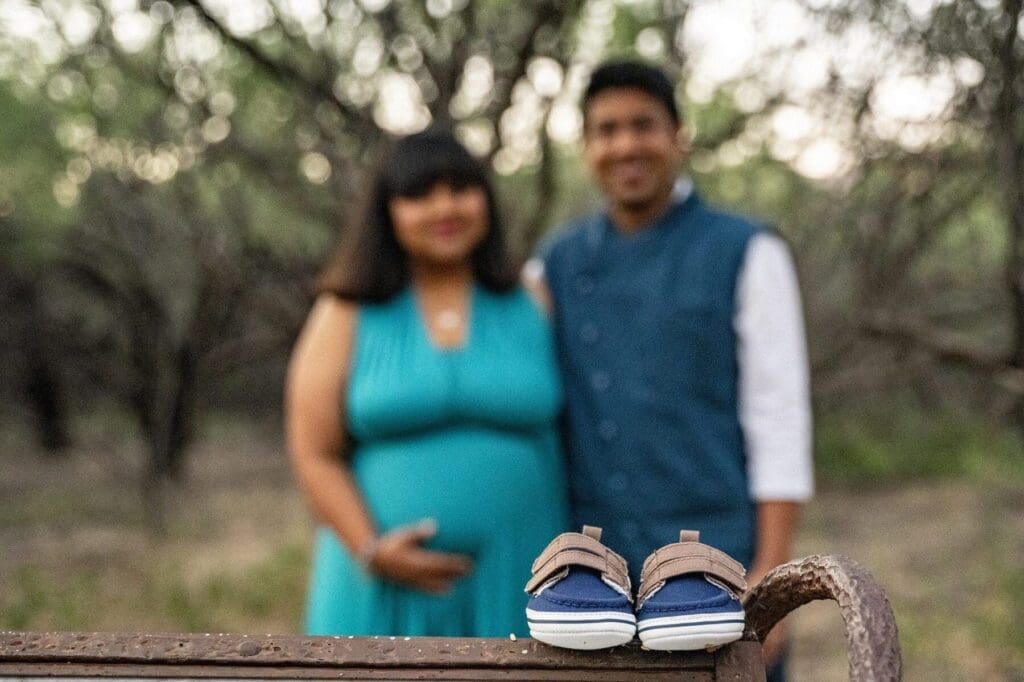 A pair of baby shoes rests in sharp focus while Aditi and her partner stand softly blurred in the wooded background.