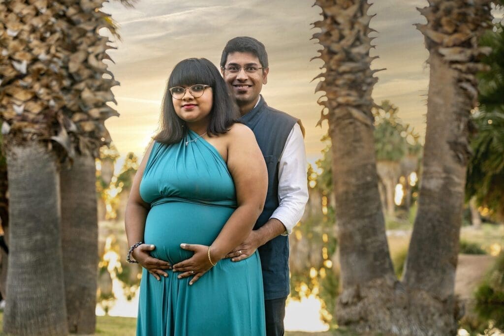 Aditi faces the camera with hands resting on her belly while her partner stands closely behind her among palm trees.