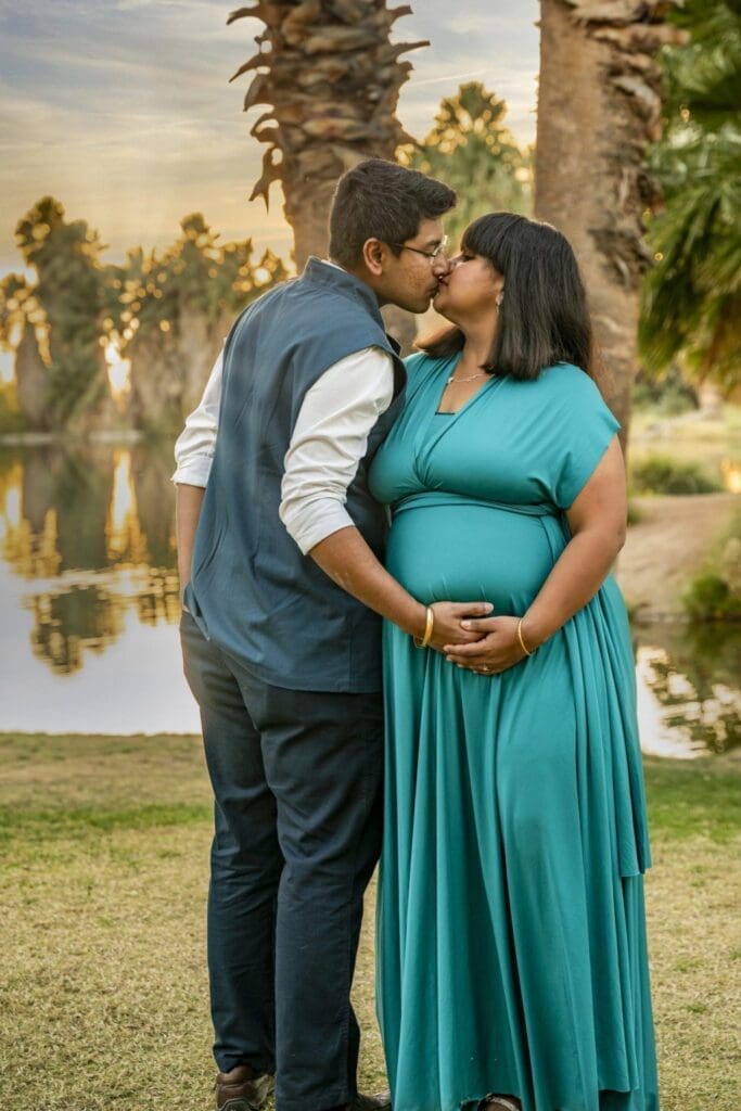 Her partner kisses Aditi as she faces the camera with both hands resting over her belly beside the water.