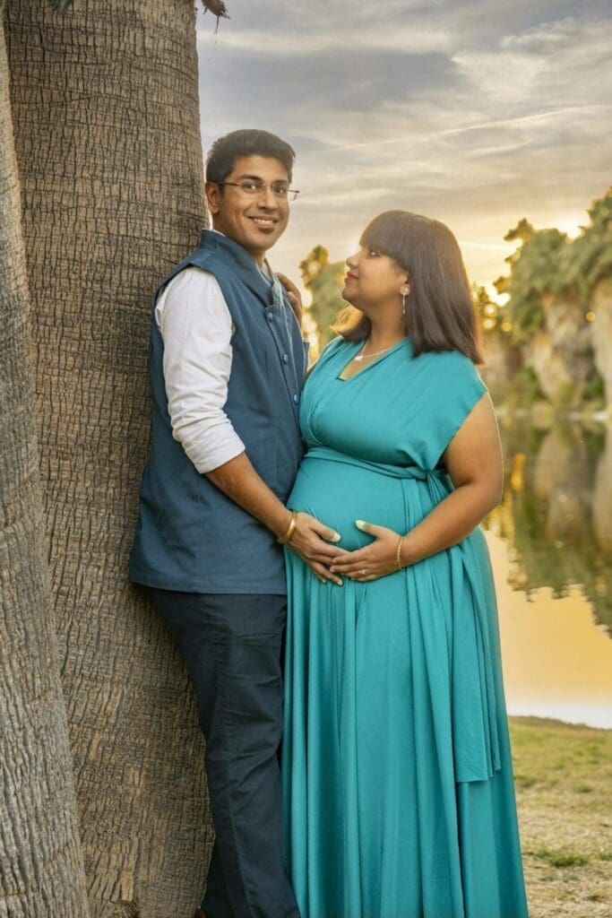 Aditi looks toward her partner while they stand closely together beside a palm trunk and pond.