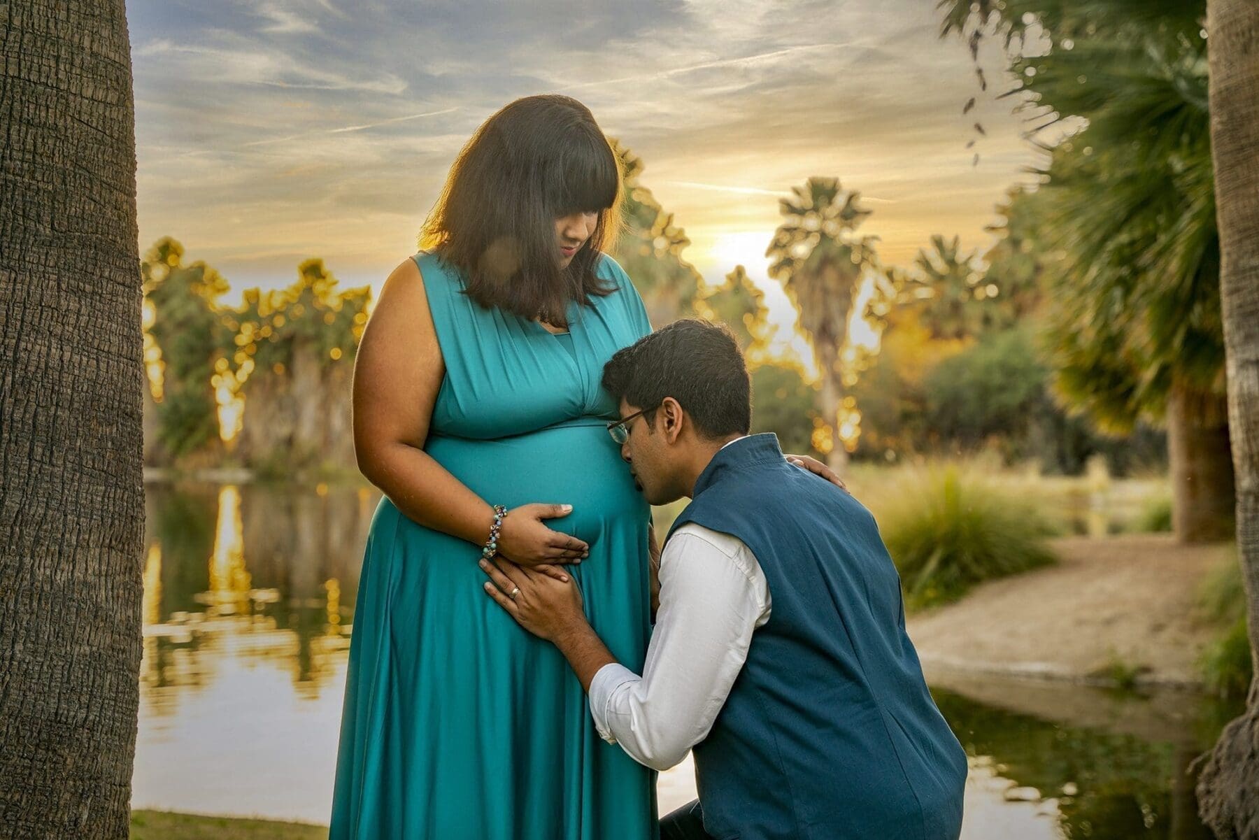 Her partner kneels in front of Aditi with hands resting gently over her belly beside a palm-lined pond.