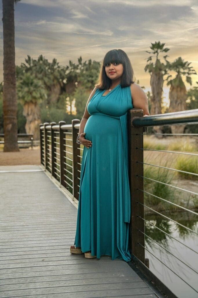 Aditi stands alone beside the bridge railing with one hand resting on her belly and palms behind her.