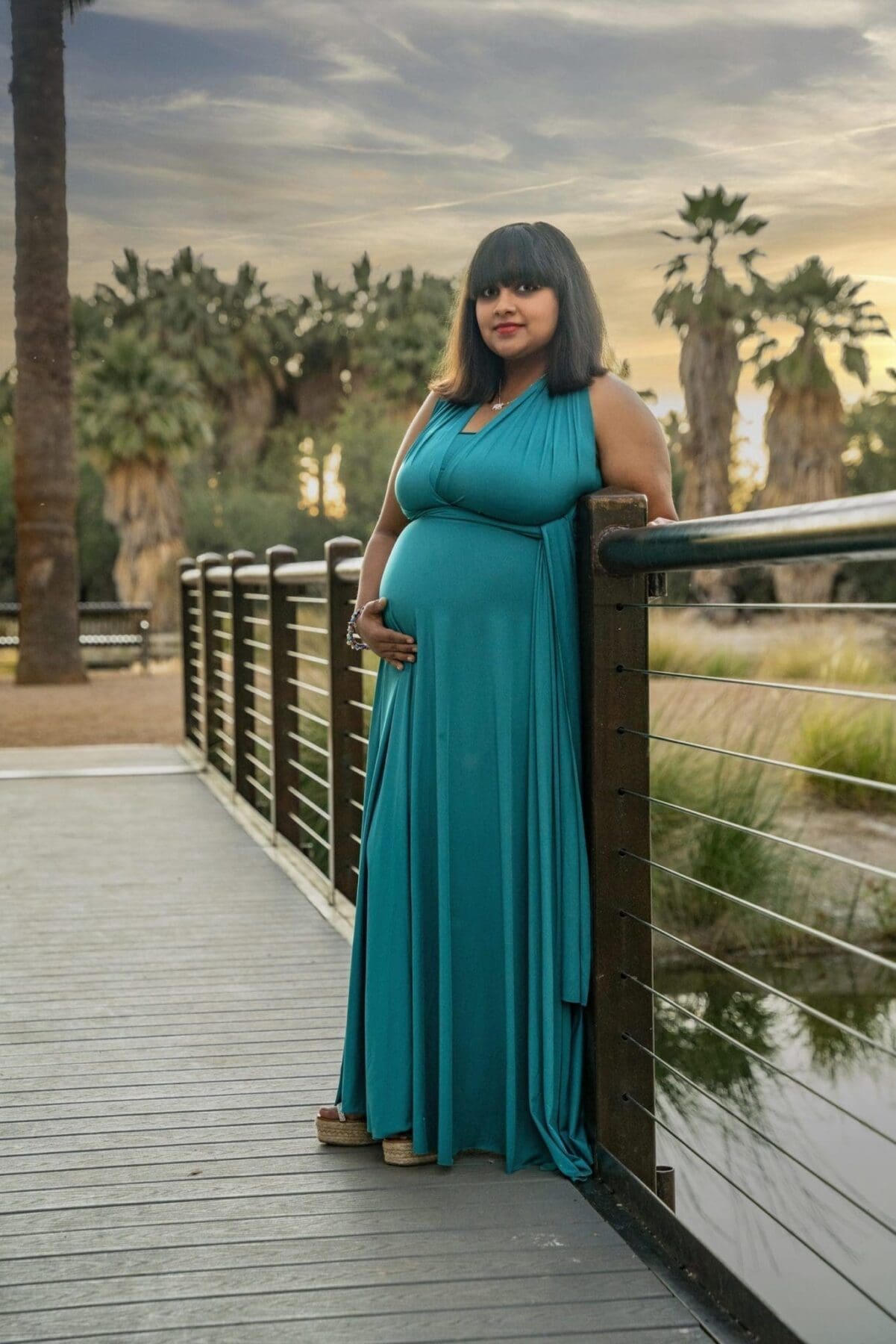 Aditi stands alone beside the bridge railing with one hand resting on her belly and palms behind her.