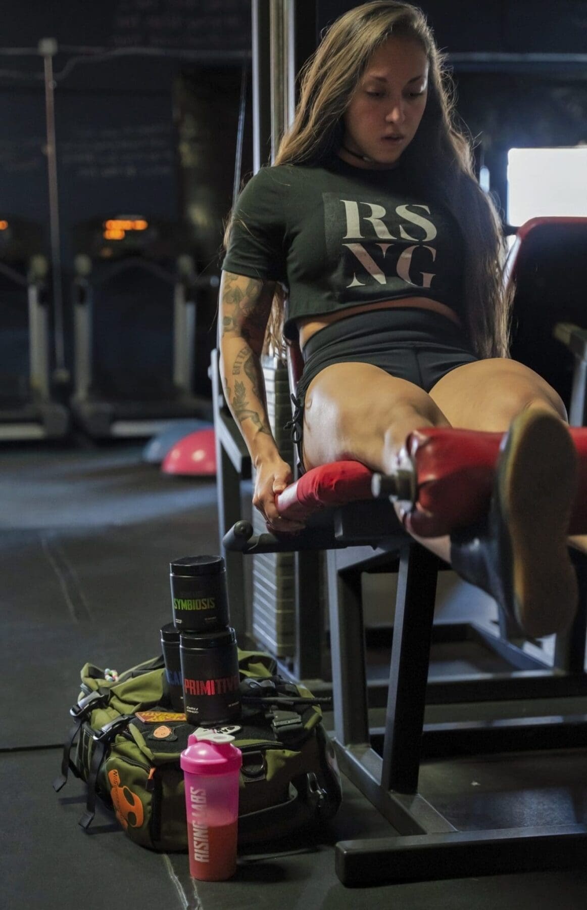 Danni performs a seated leg exercise in a dim gym with training bags and drink containers beside the machine.