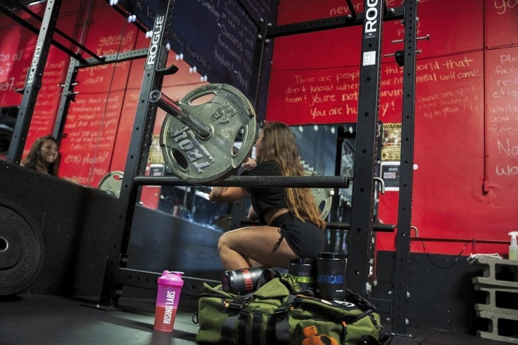 Danni performs a barbell squat inside a black power rack against a red gym wall.