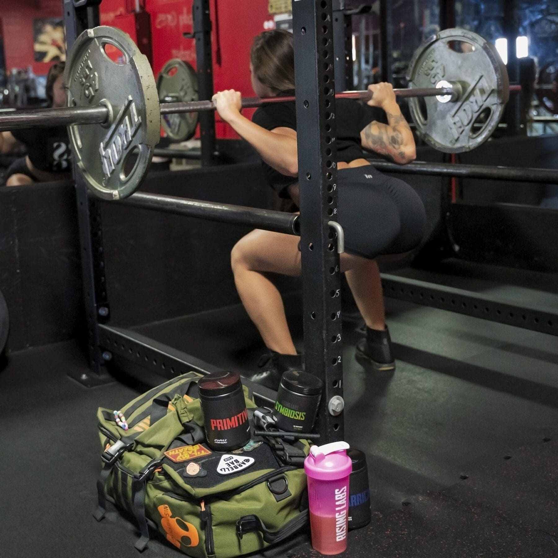 Danni holds a barbell squat between the uprights of a power rack with mirrors reflecting the gym behind her.