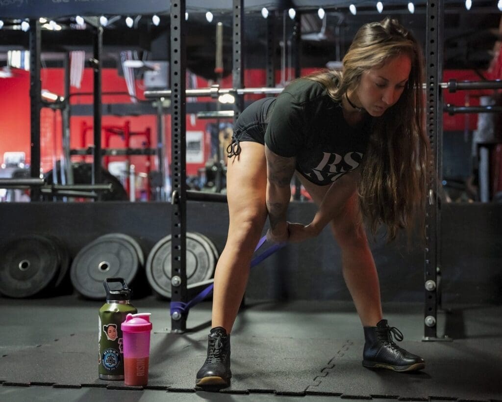 Danni bends forward in a wide stance during a floor-level resistance exercise inside the weight room.