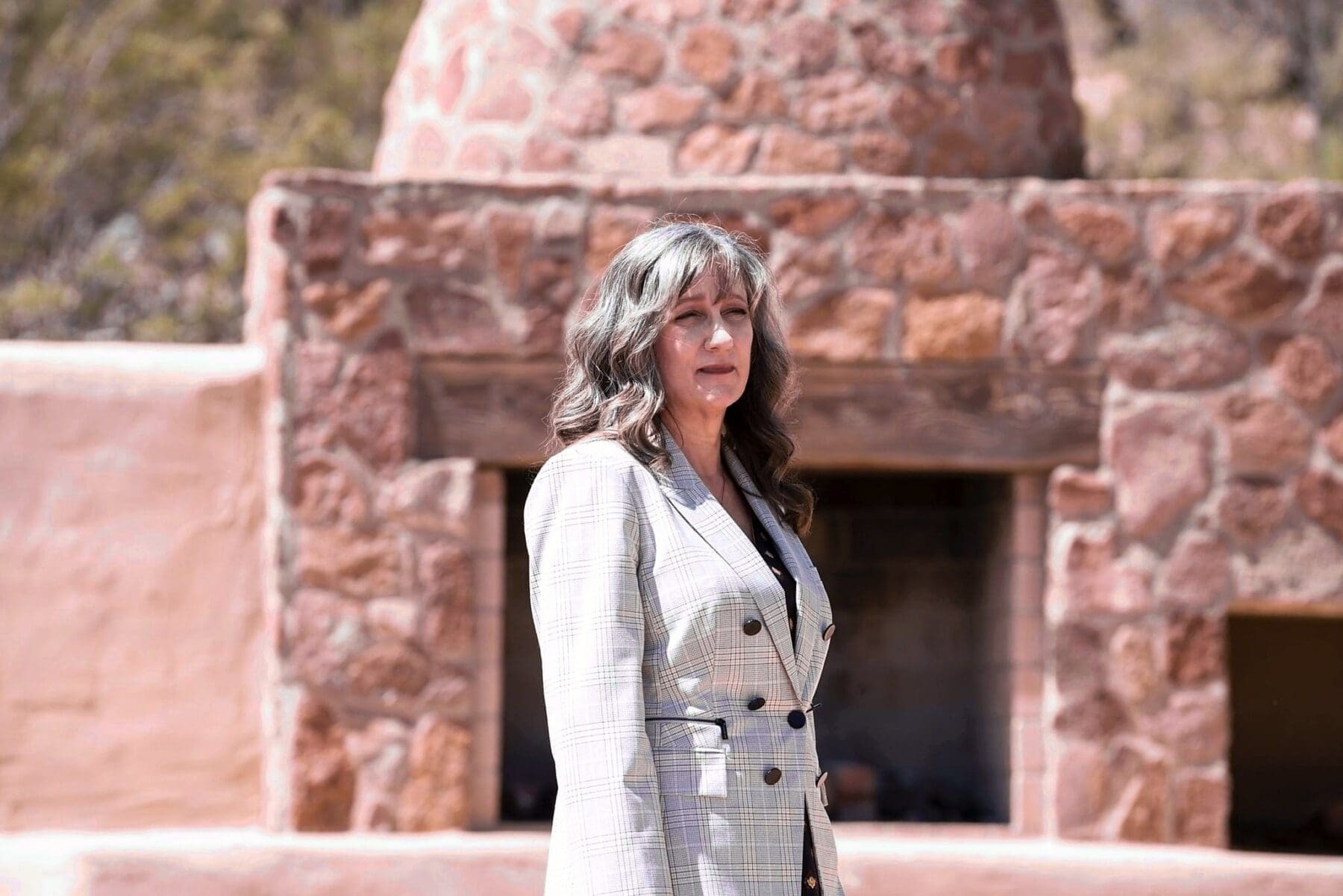 Cathy Byroad stands in a light plaid blazer in front of a rustic outdoor stone fireplace.