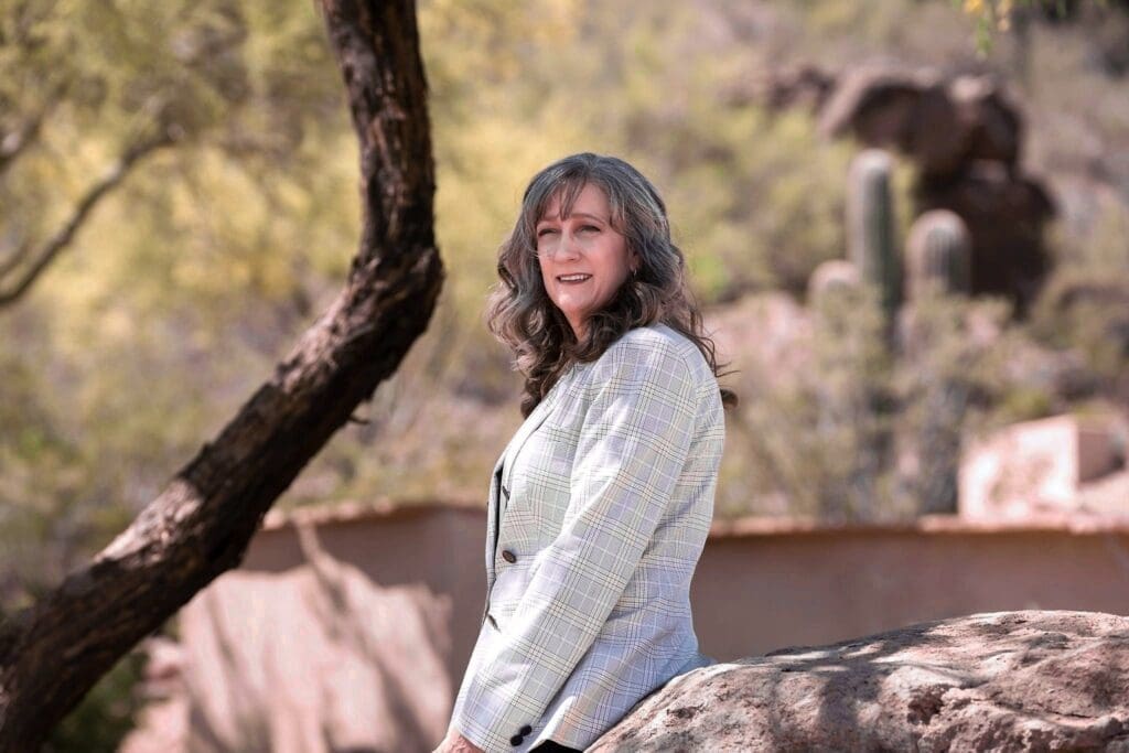 Cathy Byroad smiles beside a large boulder with desert trees and cactus behind her.