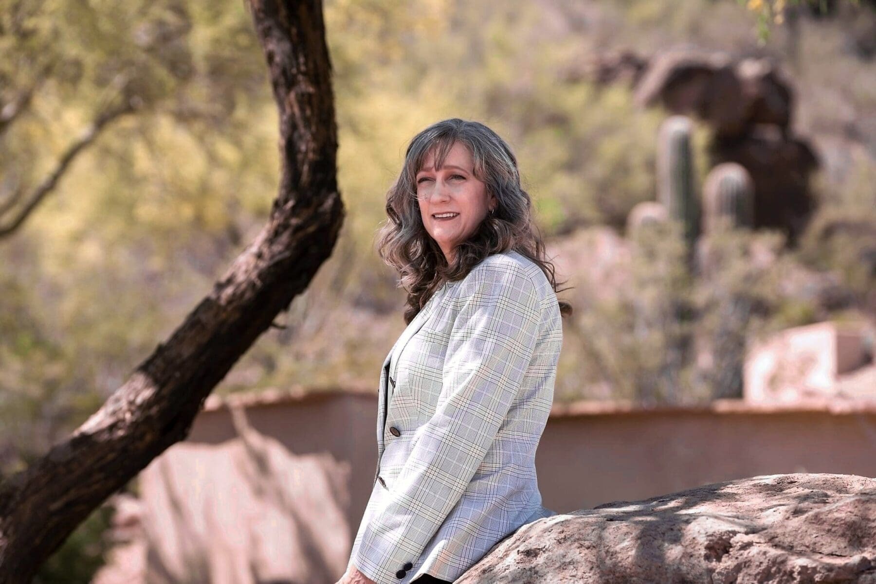 Cathy Byroad smiles beside a large boulder with desert trees and cactus behind her.