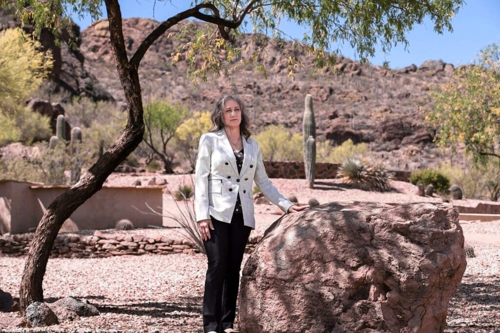 Cathy Byroad stands beside a boulder in a broad desert courtyard framed by trees and saguaros.
