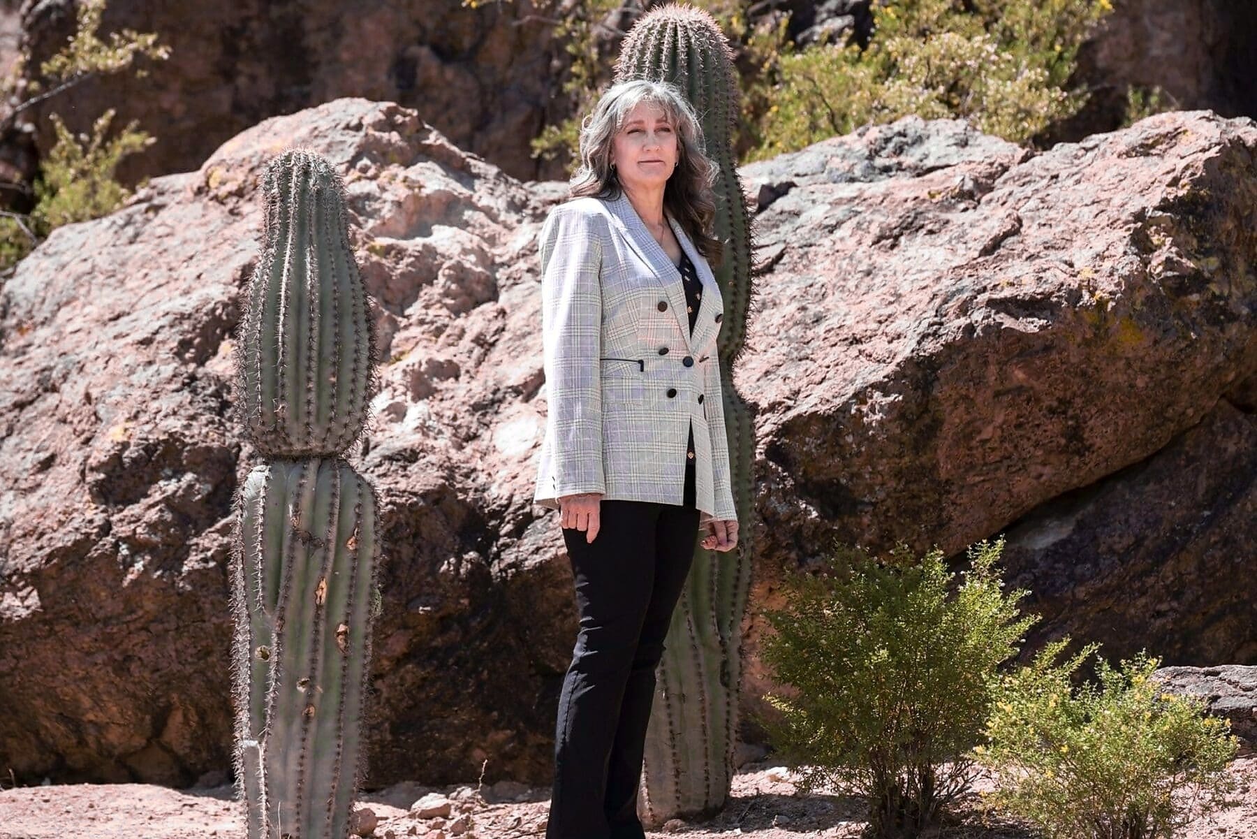 Cathy Byroad stands beside a tall cactus and sunlit desert boulders.