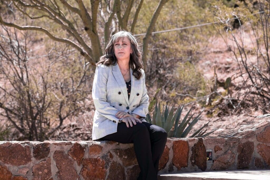Cathy Byroad sits on a low stone wall beneath a desert tree.