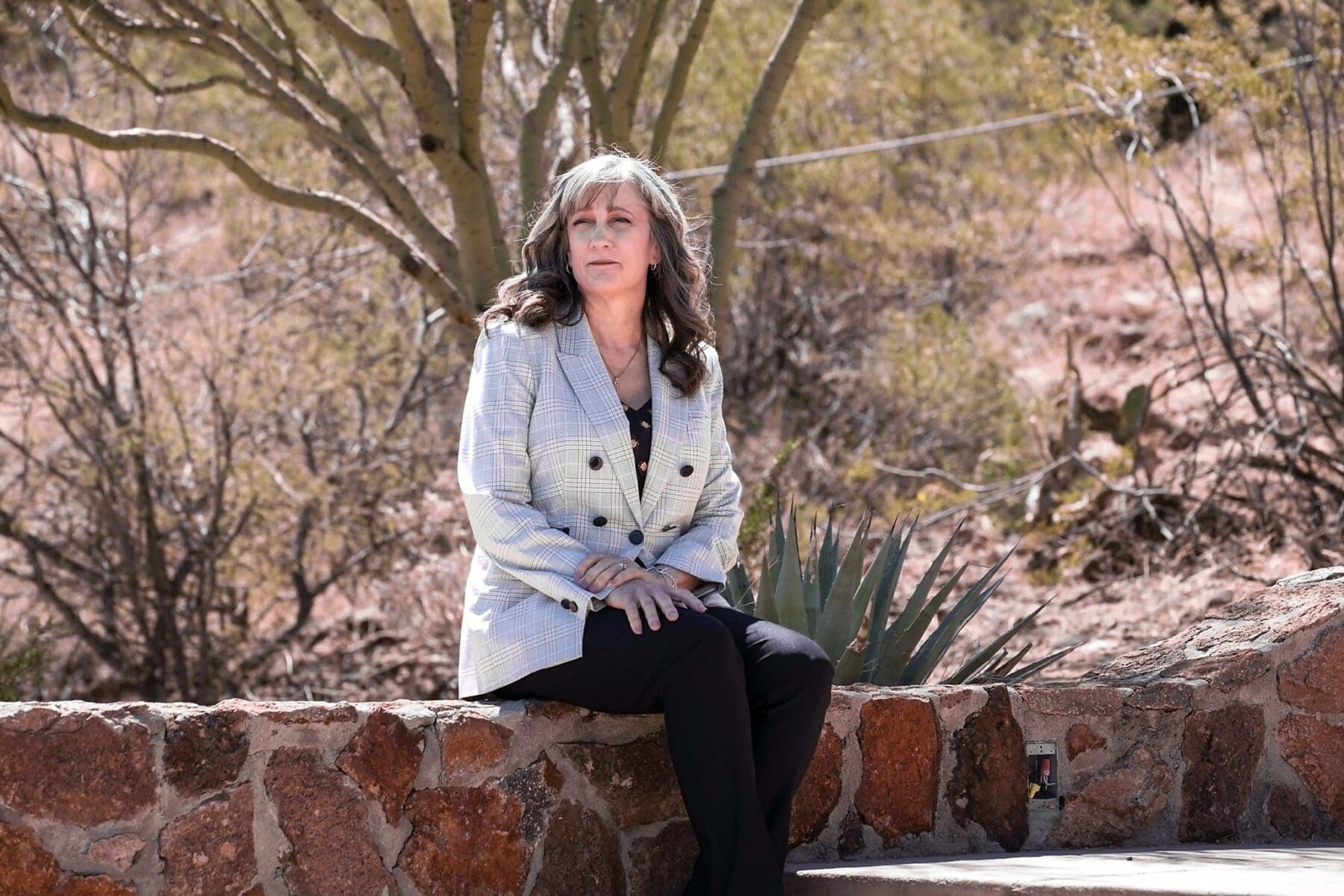 Cathy Byroad sits on a low stone wall beneath a desert tree.