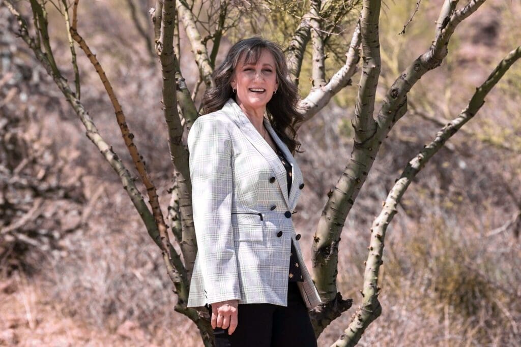 Cathy Byroad smiles beside pale desert tree trunks in a light plaid blazer.