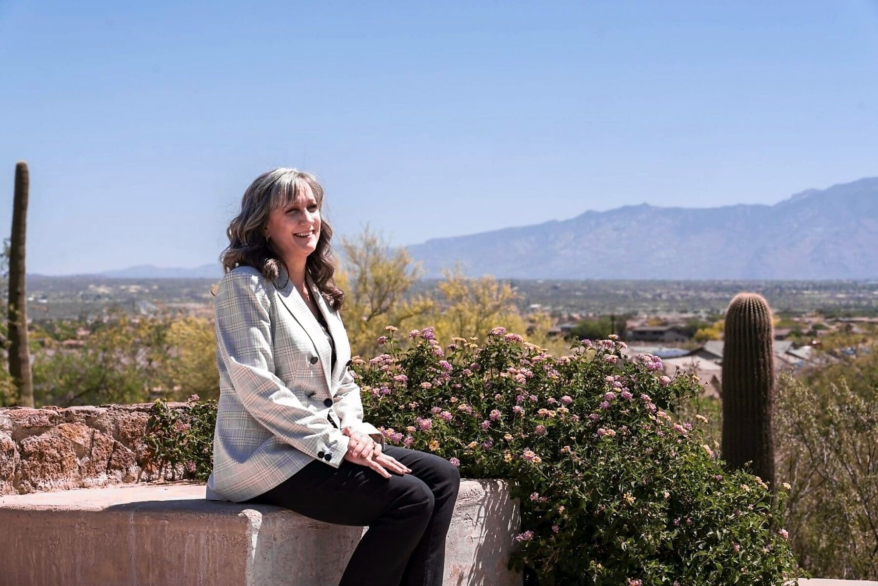 Cathy Byroad sits on a low wall with flowering shrubs, saguaros, and distant mountains behind her.