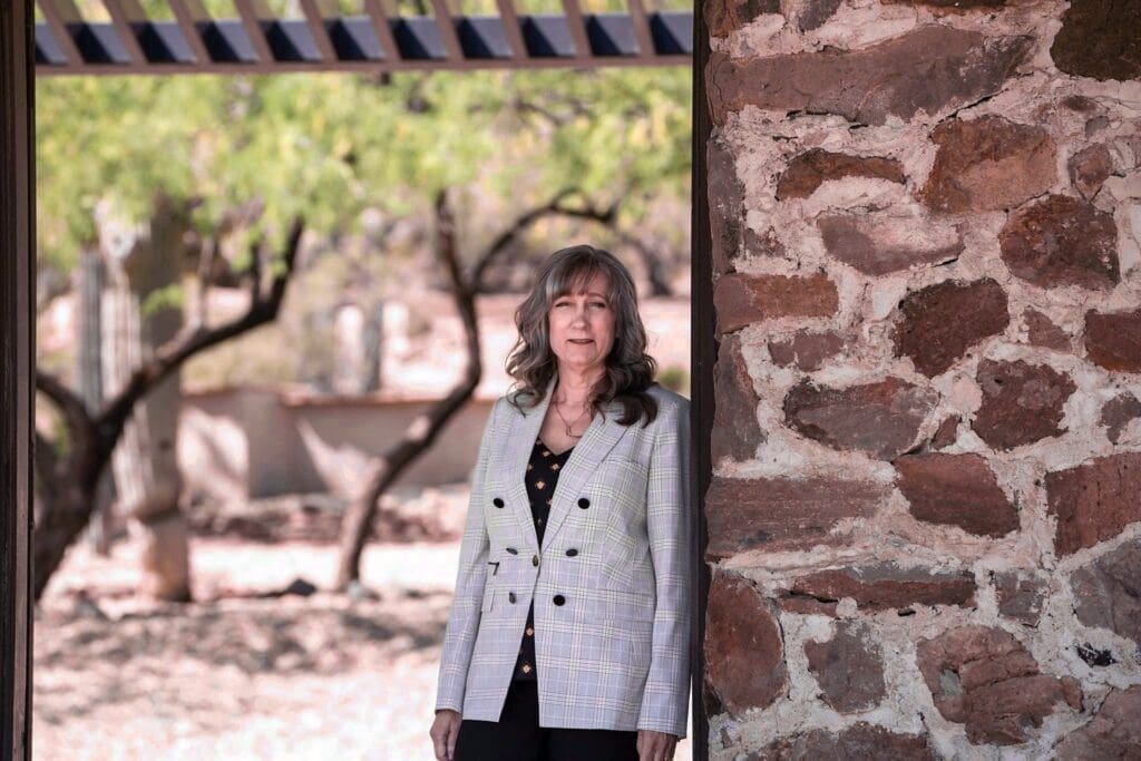 Cathy Byroad stands beside a rustic stone wall with a shaded courtyard behind her.