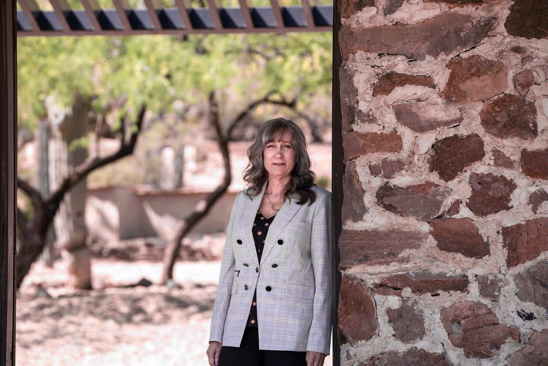 Cathy Byroad stands beside a rustic stone wall with a shaded courtyard behind her.