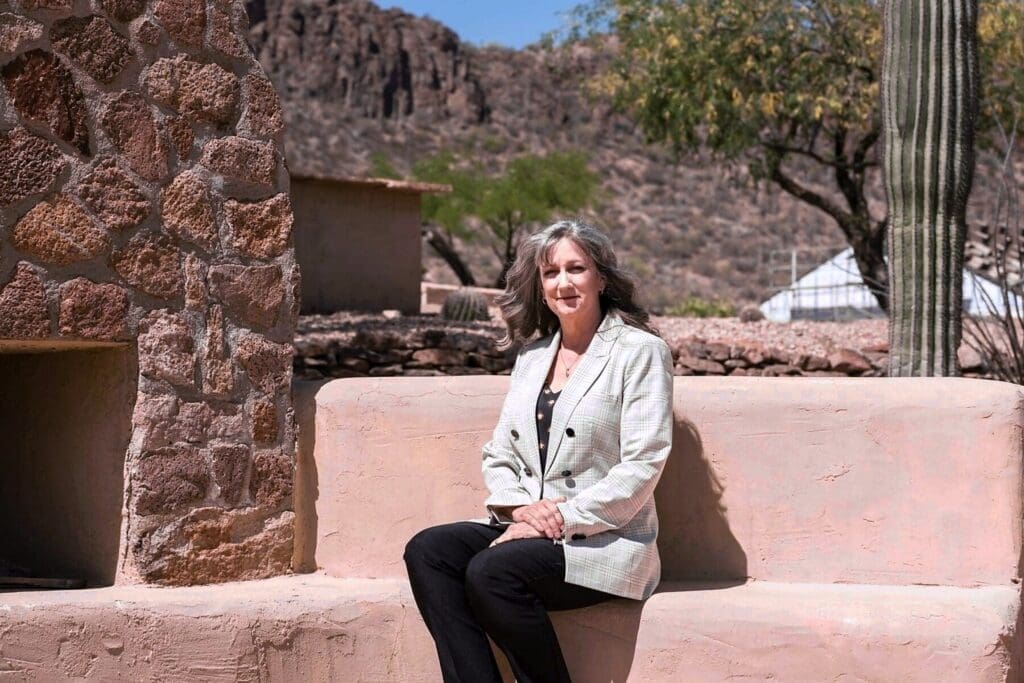 Cathy Byroad sits on an adobe bench beside a stone fireplace and saguaro.