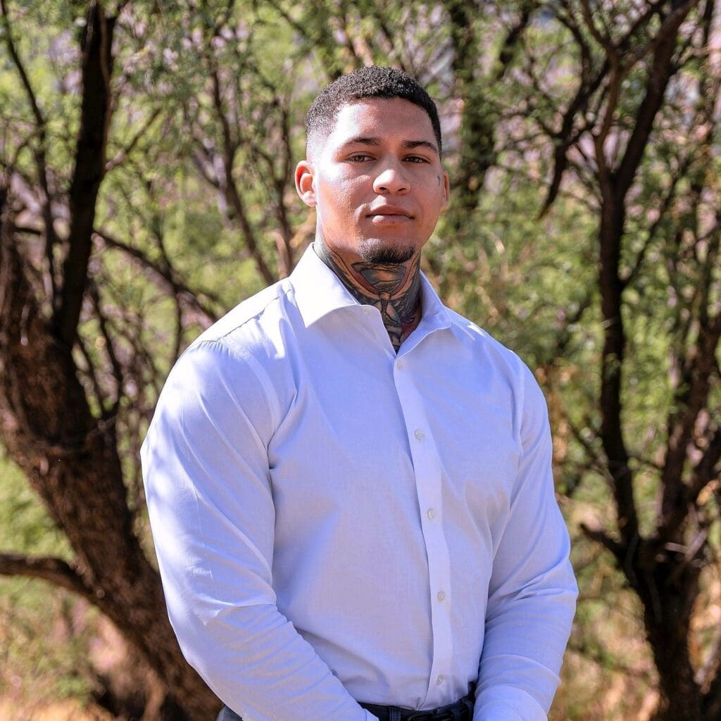A young man in a white shirt stands in front of softly blurred desert trees.