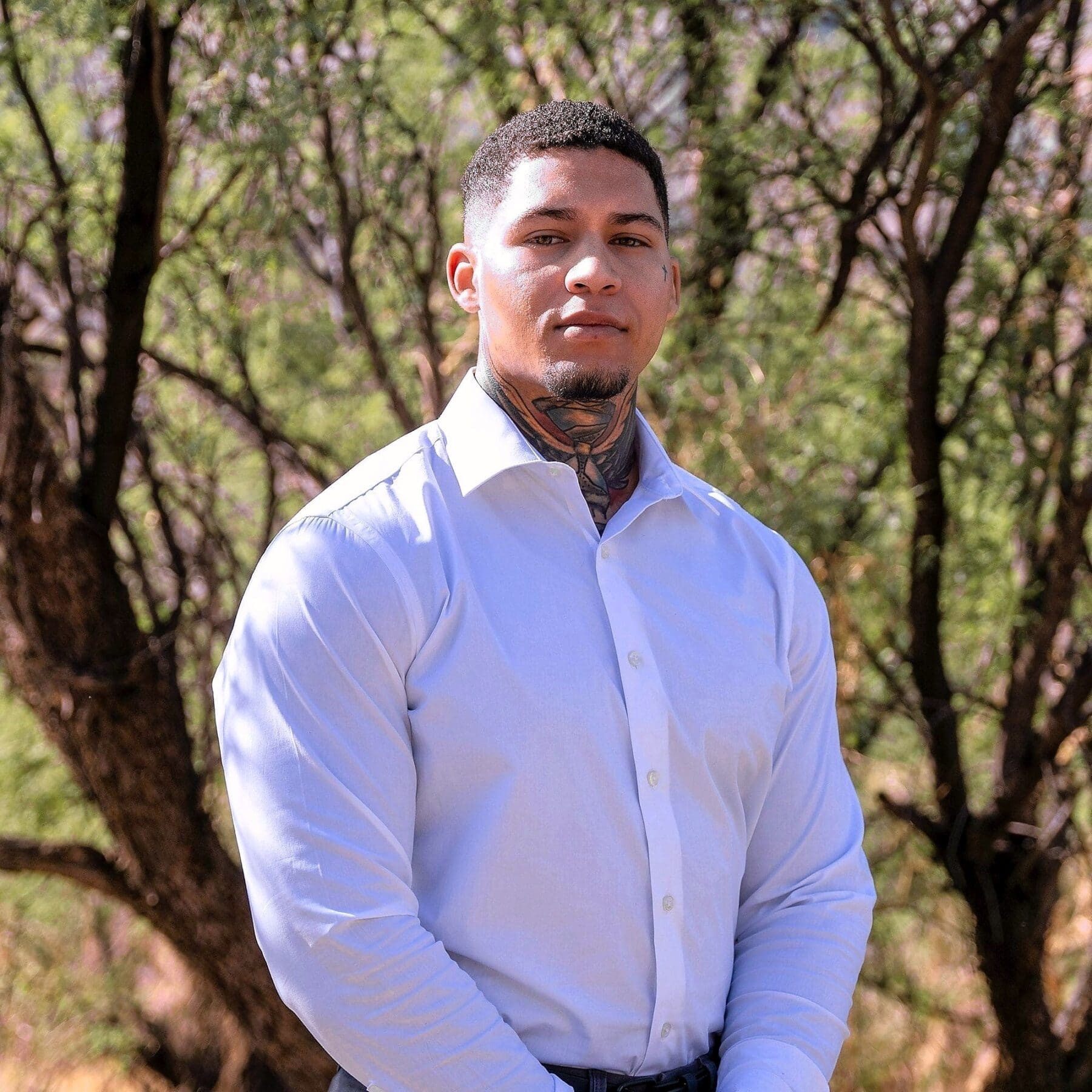 A young man in a white shirt stands in front of softly blurred desert trees.