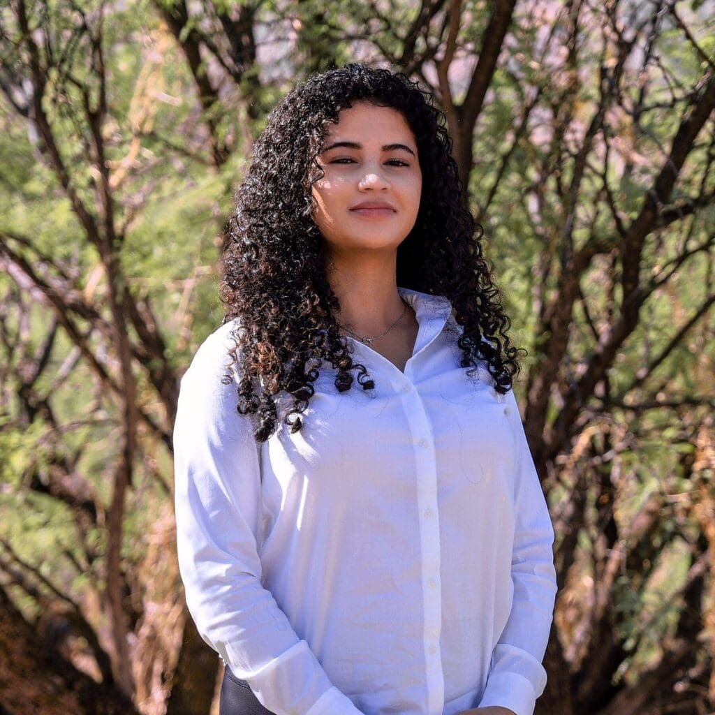 A young woman with long curly hair poses in a close portrait among desert trees.