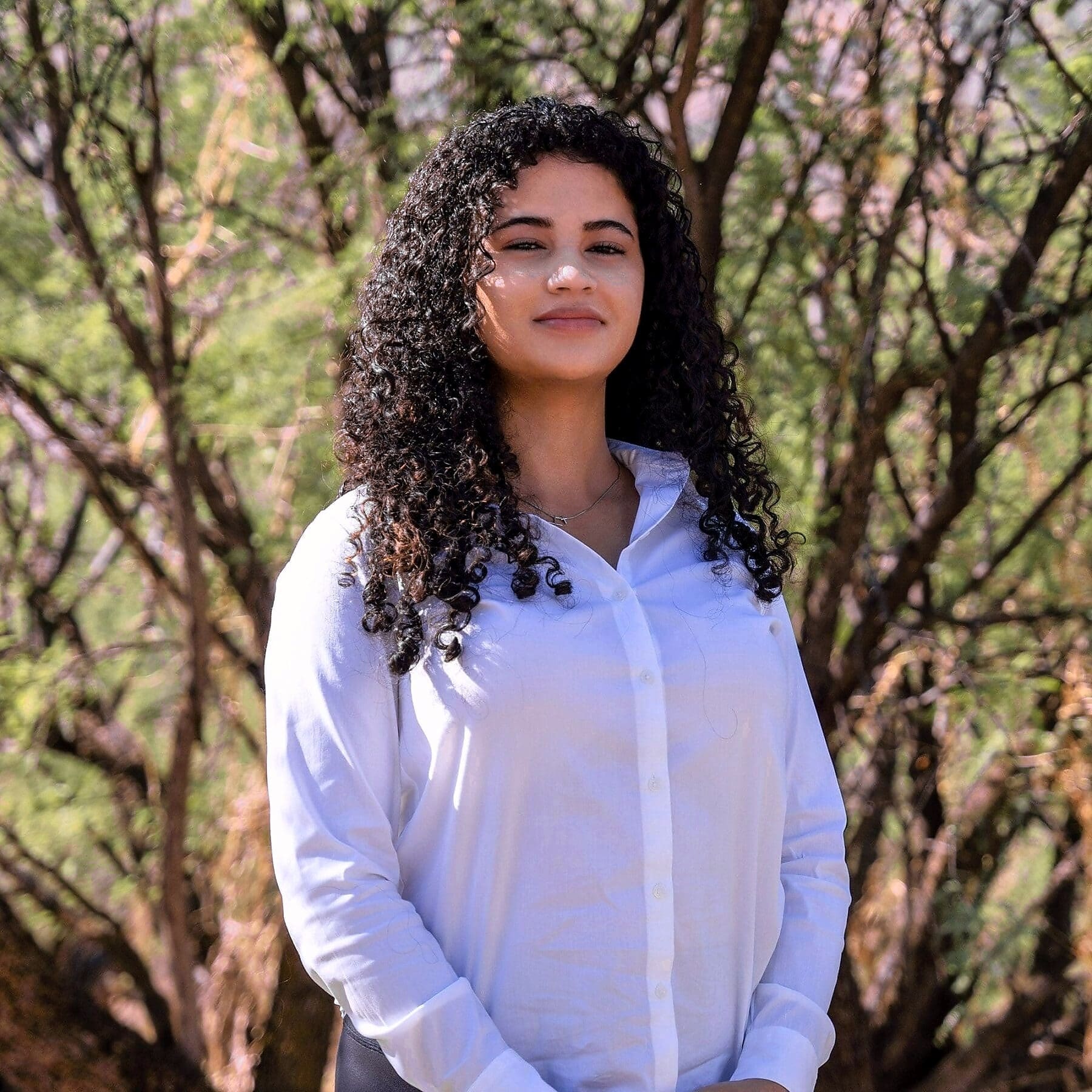 A young woman with long curly hair poses in a close portrait among desert trees.