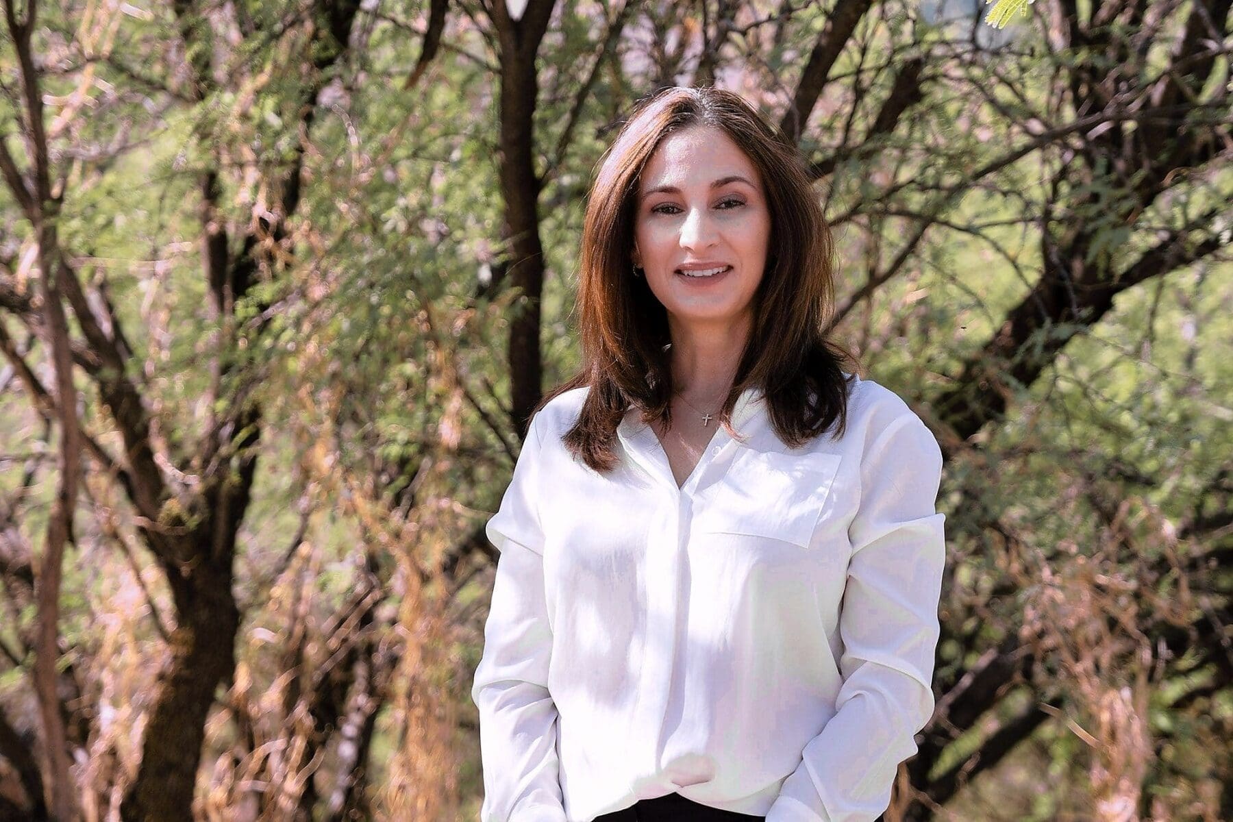 A woman in a white blouse faces the camera in a close portrait among desert greenery.