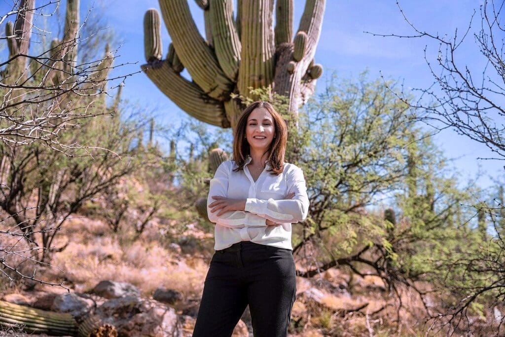 A woman in a white blouse stands with folded arms beneath a large saguaro.