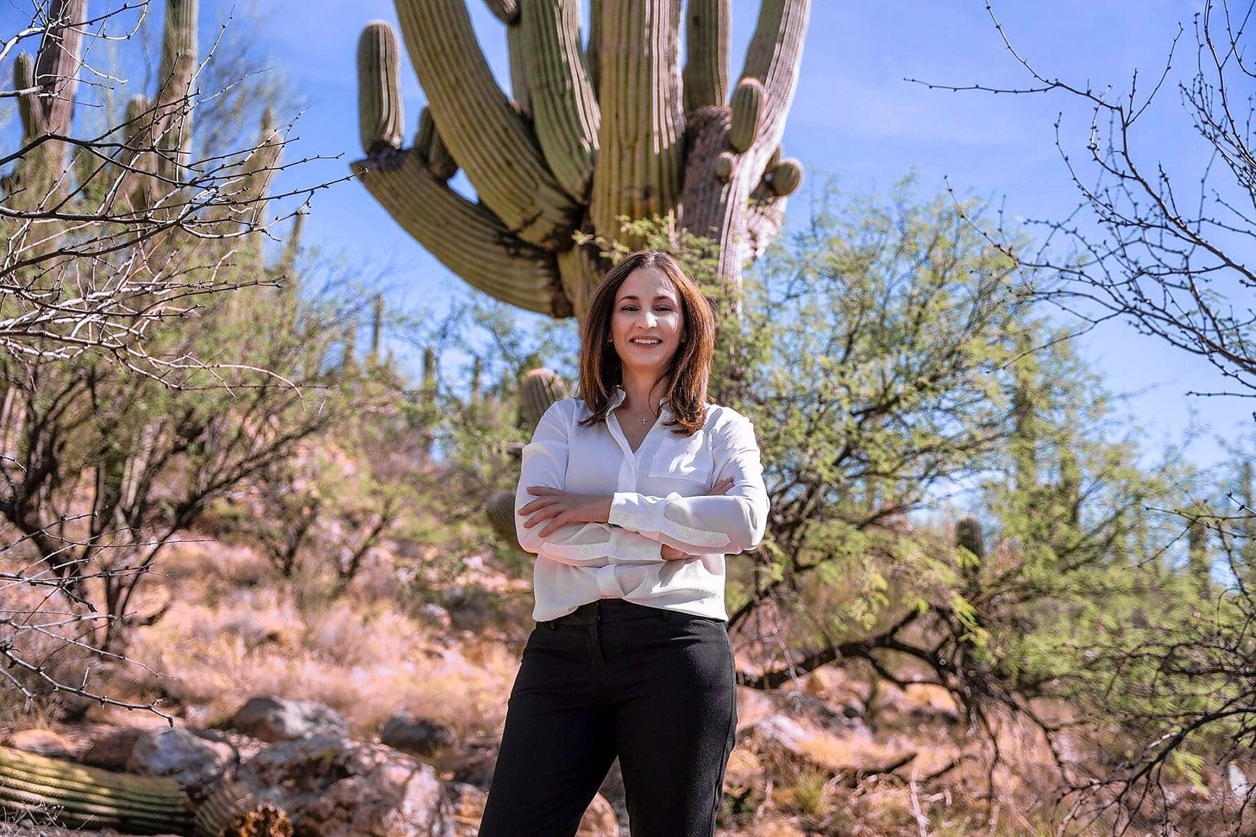 A woman in a white blouse stands with folded arms beneath a large saguaro.