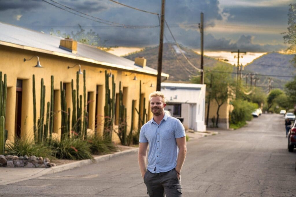 Chris Romero stands in a narrow street beneath dramatic sunset clouds with mountains in the distance.