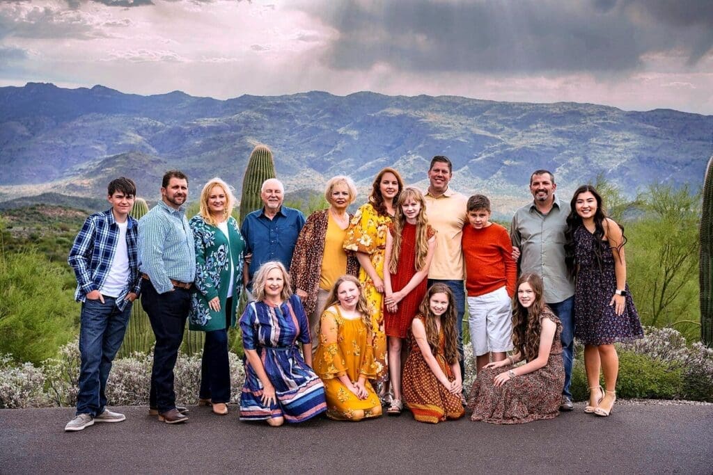 A large family group poses beside a saguaro with a broad mountain range and dramatic clouds behind them.