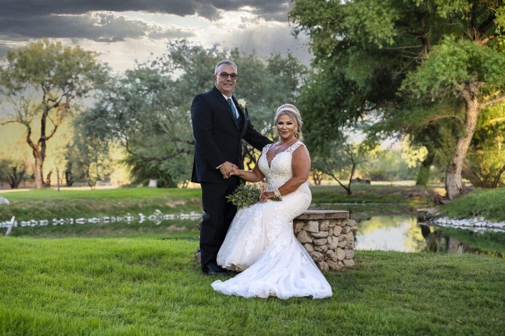 The wedding couple stands together on the lawn beside a low stone wall and pond.