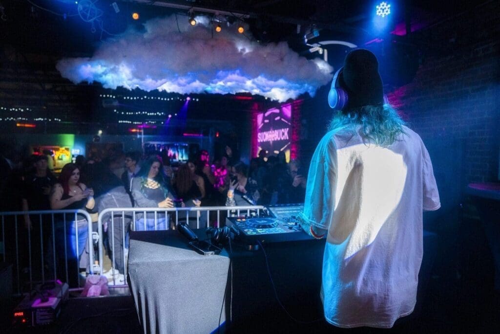 An over-the-shoulder view places the DJ, mixing console, and audience together beneath blue light.