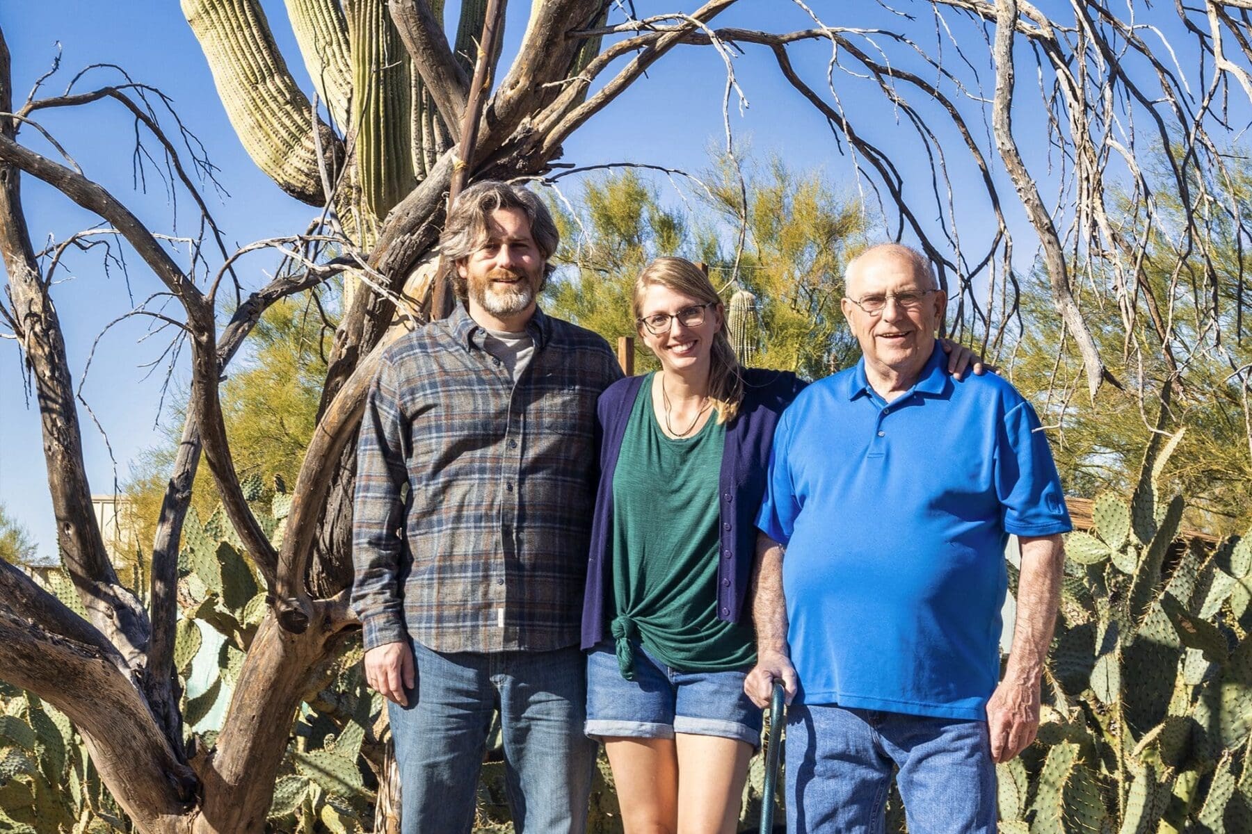 Three adults stand together beneath a leafless desert tree with cactus and blue sky behind them.