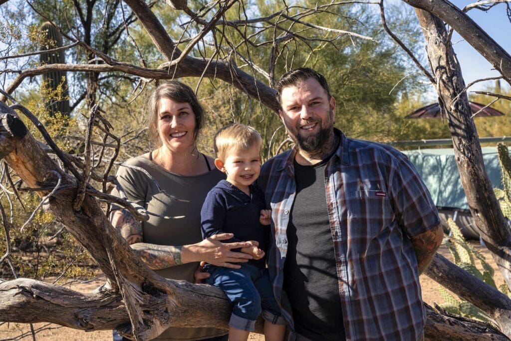 Two adults and a young child pose together beside trees and desert plants.