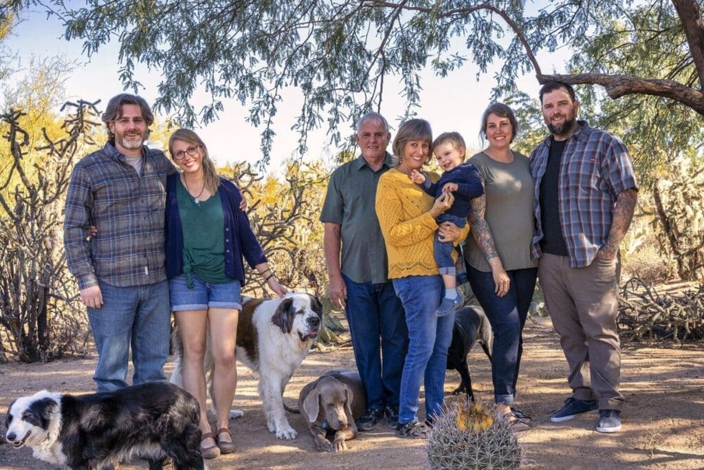 A large group of adults and a young child pose on a desert path with several dogs.