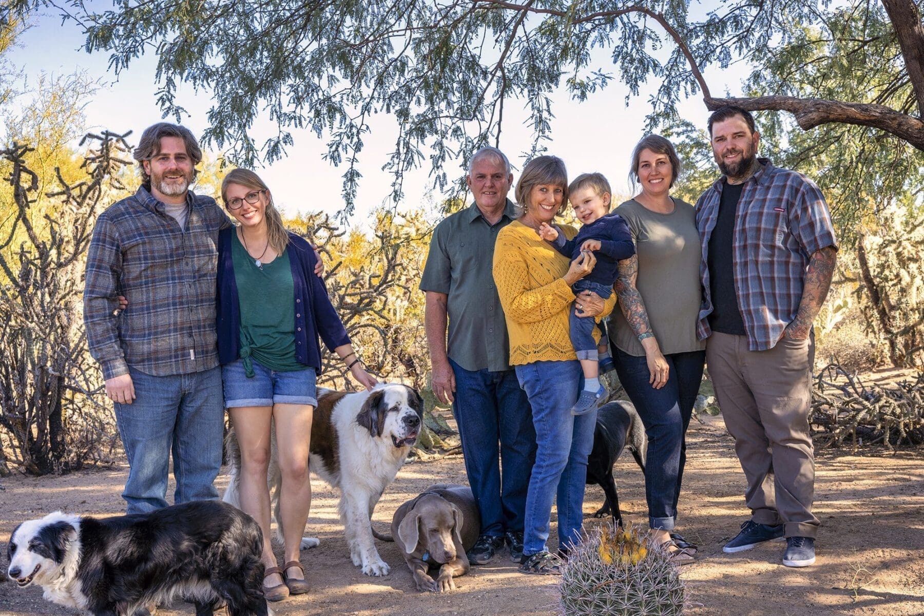 A large group of adults and a young child pose on a desert path with several dogs.