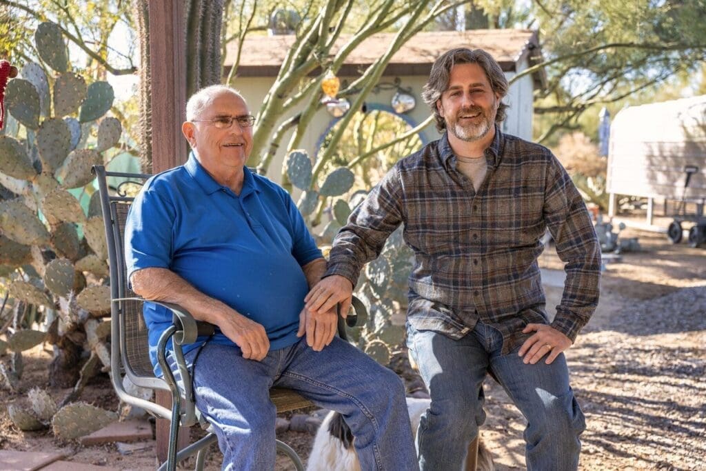 Two men sit side by side in a desert yard with cactus behind them.